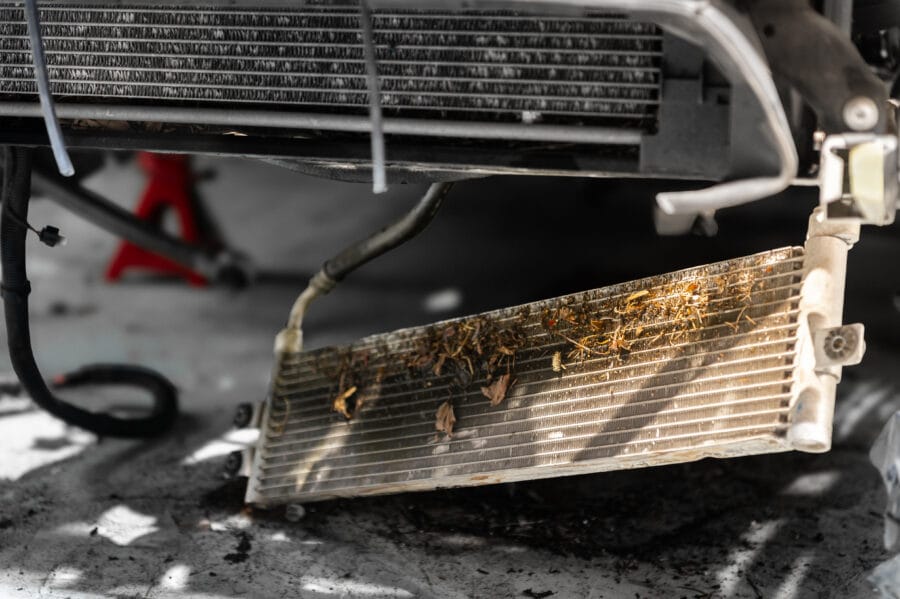 Underside view of a BMW F82's OEM oil cooler core, packed with leaves, dirt, and debris blocking the fins, seen next to another cooler radiator above it.