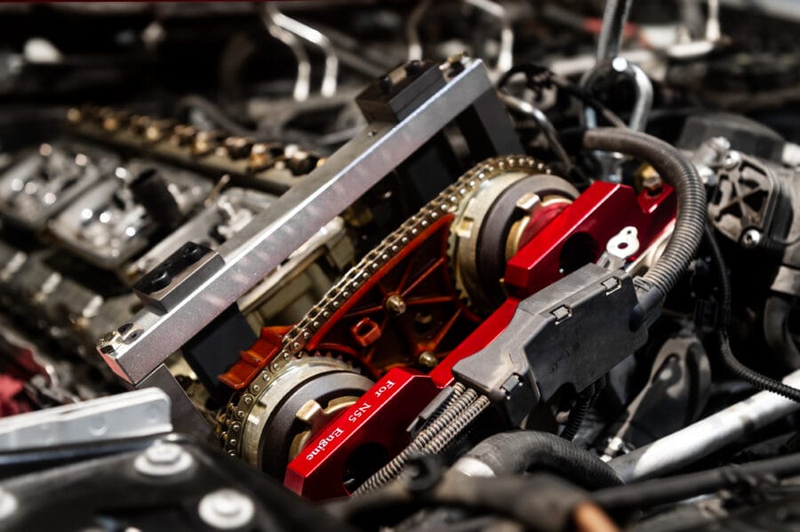 Close-up of a BMW S55 engine's timing chain and gears with a red billet timing tool installed, surrounded by engine components in the bay.