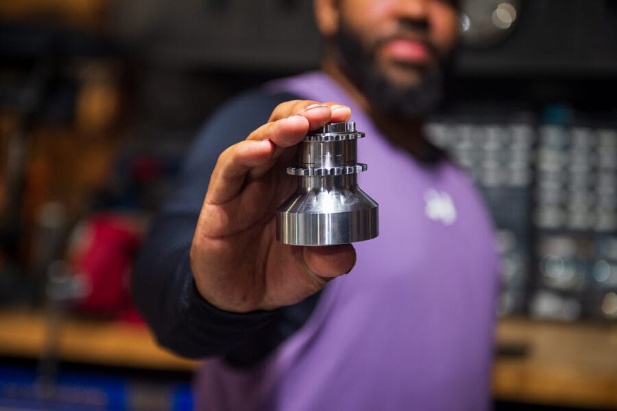 Devin Giles holding a machined aluminum S55 pinned crank hub up to the camera in a workshop before installation.