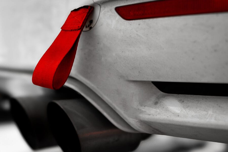 Close-up of a red rear tow strap with a cut slit for a cleaner look, mounted below the taillight of a white BMW F82 M4 above the dual exhaust tips.