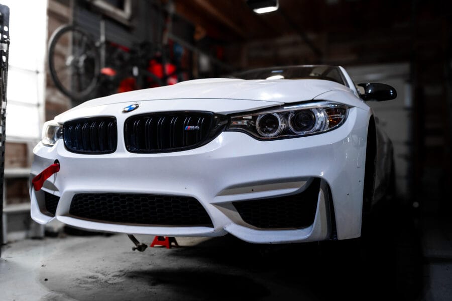 Close-up front-end view of a white BMW F82 M4 with black kidney grilles and headlights, raised on jackstands in a home garage.