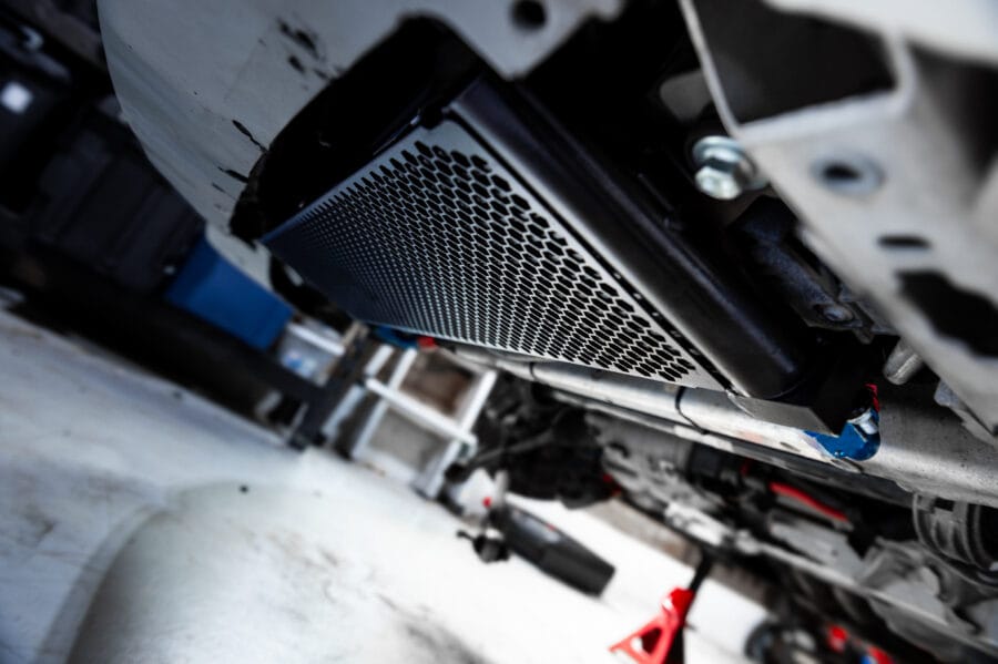 Close-up underside view of a CSF oil cooler mounted behind the front bumper of a BMW F82 M4, with mesh cooler core and surrounding suspension components visible.