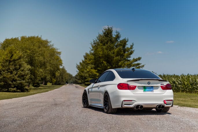 White BMW F82 M4 with black wheels and carbon trunk spoiler, rear three-quarter view parked on a gravel country road lined with trees and cornfields under a clear blue sky.