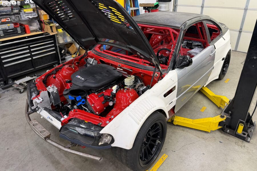 White and bare-metal BMW E46 M3 GTR clone with hood open, showing red-painted engine bay and roll cage, parked on a lift in a garage during a Vorshlag build for the Ultimate Track Car Challenge.