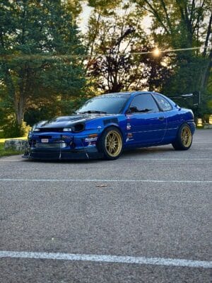 Blue 1999 Dodge Neon race car with a front splitter, support rods, fender louvers, and rear wing