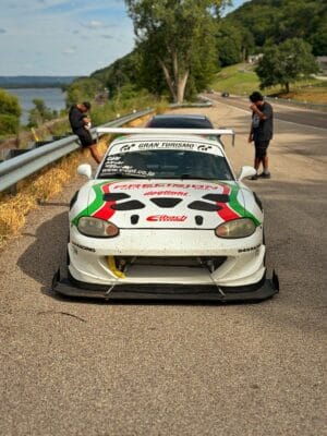 Front view of Joseph Herr’s white 2000 Mazda Miata NB with a quick-release front splitter, support rods, and titanium protection hardware