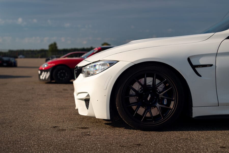 Front-side view of a white BMW F82 M4 fitted with black Apex wheels and Paragon two-piece brake rotors, parked in a paddock with other cars in the background.