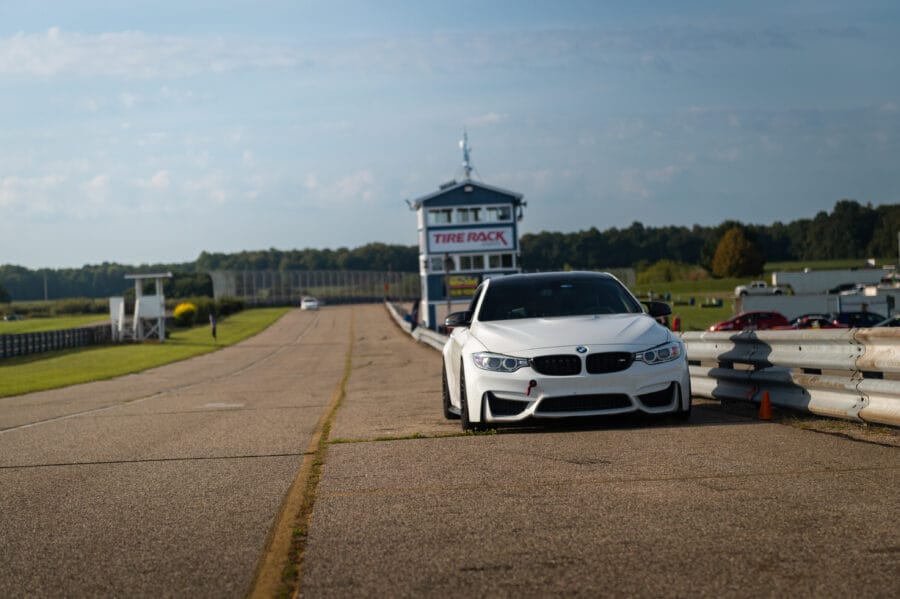 White BMW F82 M4 with black wheels parked in the pit lane at Gingerman Raceway, with the Tire Rack timing tower and track visible in the background.