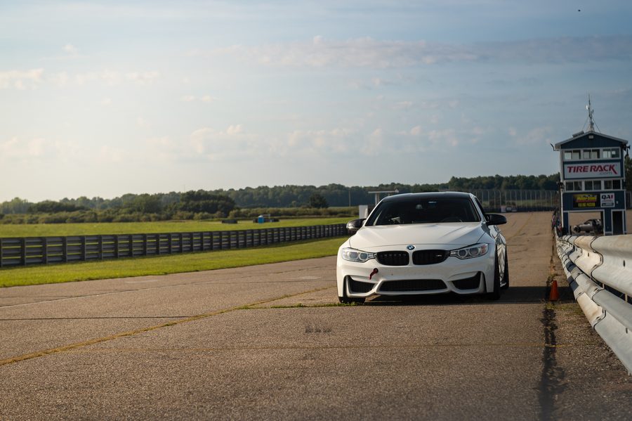 White BMW F82 M4 parked on pit lane at Gingerman Raceway with the Tire Rack timing tower and track fencing visible in the background.