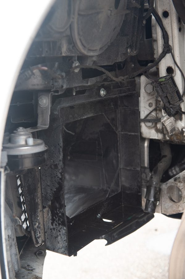 Bare BMW F82 M4 front corner with the splash shield removed and radiator missing, showing the empty plastic radiator cage and surrounding wiring harness