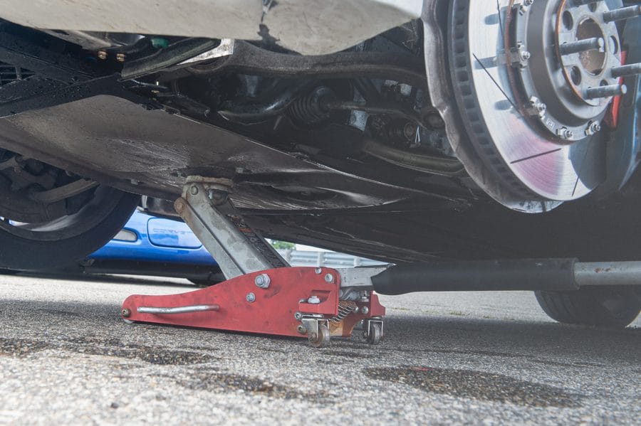 Low-angle view of a red aluminum track jack lifting the BMW F82 M4, showing a wet undercarriage, brake rotor, and steering components after a coolant leak.