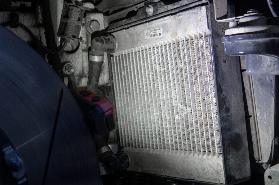 Backside view of a BMW F82 M4's auxiliary cooler with visible BEHR part label, showing coolant hoses, fittings, and windshield washer reservoir nearby.