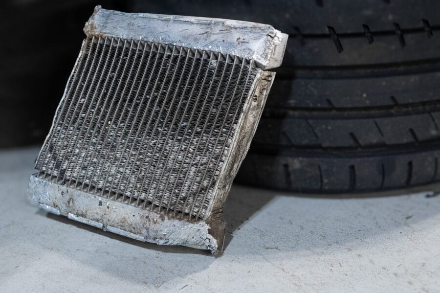 A removed BMW F82 M4 auxiliary cooler with a torn, corroded end tank, leaning against a tire on a garage floor.