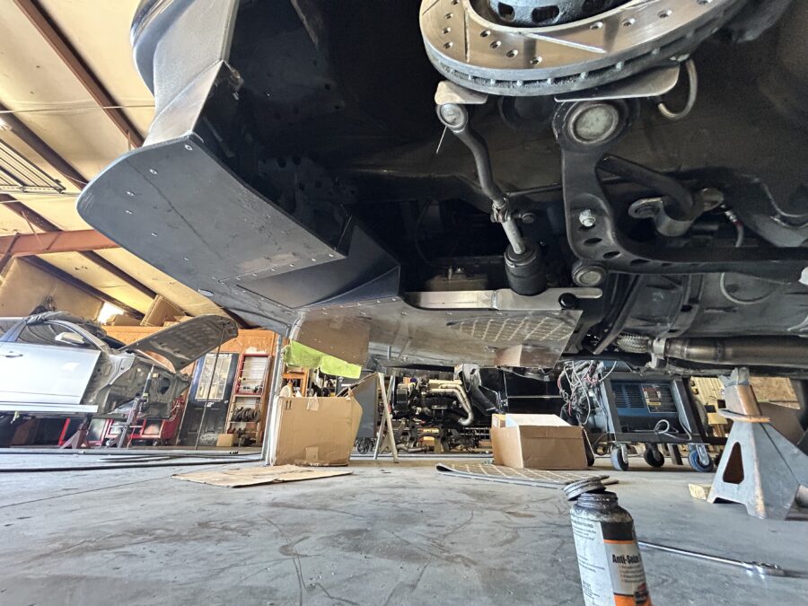 Underbody view of a BMW E30 showing a splitter diffuser, aluminum air dam, and surrounding suspension components.