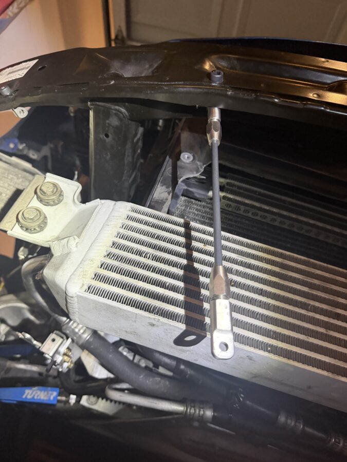 Close-up of a splitter support rod bolted through the OEM front bumper bar on Tanner’s BMW E82.