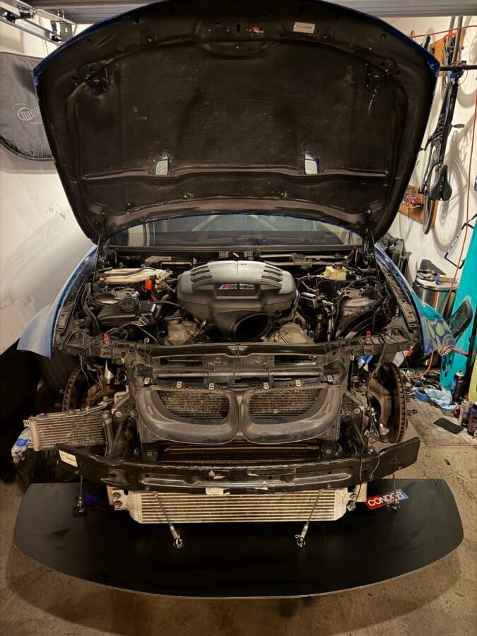 Front view of Tanner’s BMW E82 with the bumper removed, showing the S65 engine, front splitter, and splitter support rods.